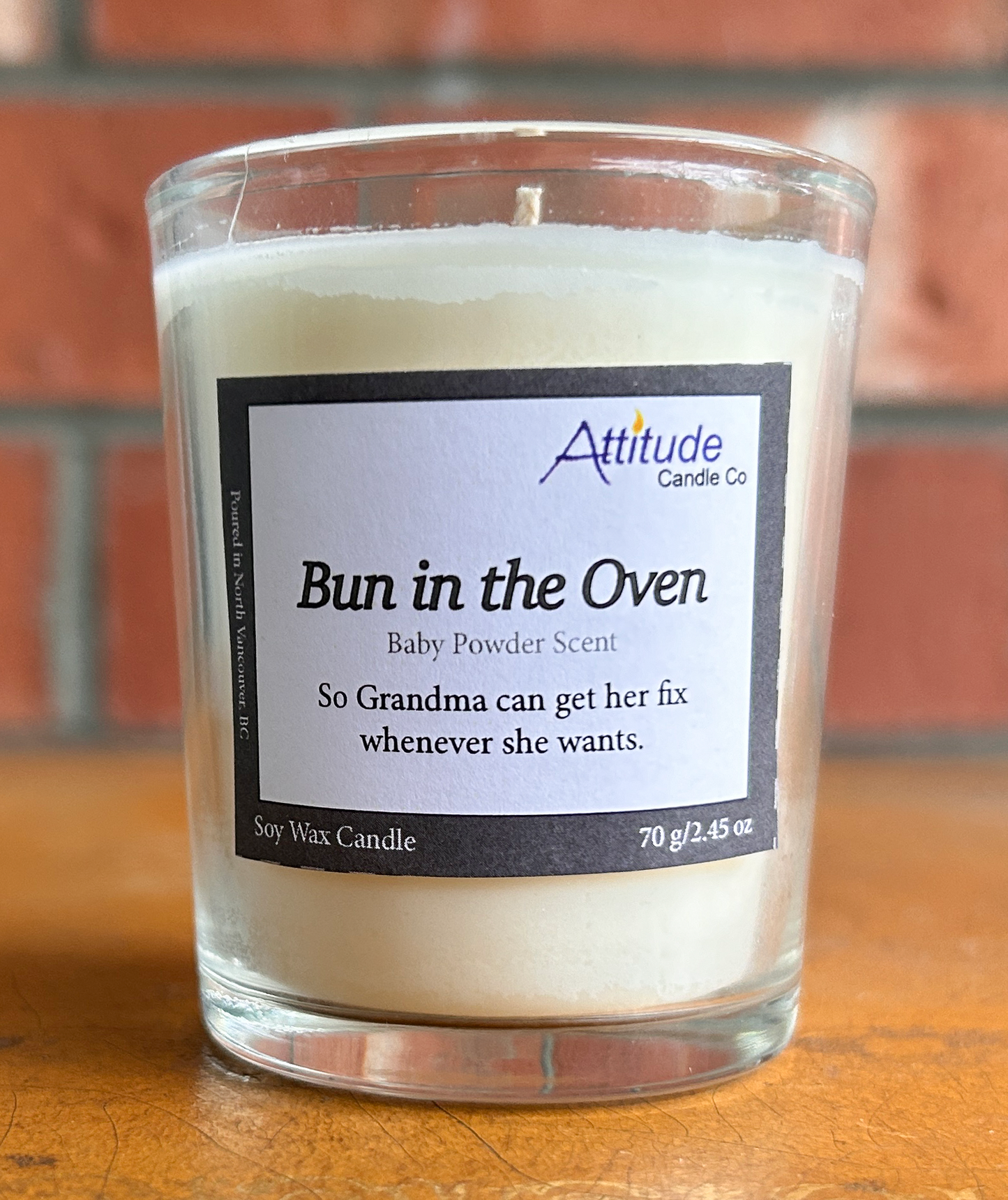 A picture of a small votive candle in a jar. The label says "Bun in the Oven", "Baby Powder Scent", and "So Grandma can get her fix whenever she wants". The label has an Attitude Candle Co logo in the top right corner. The candle is sitting on a brown table with a brick background behind.