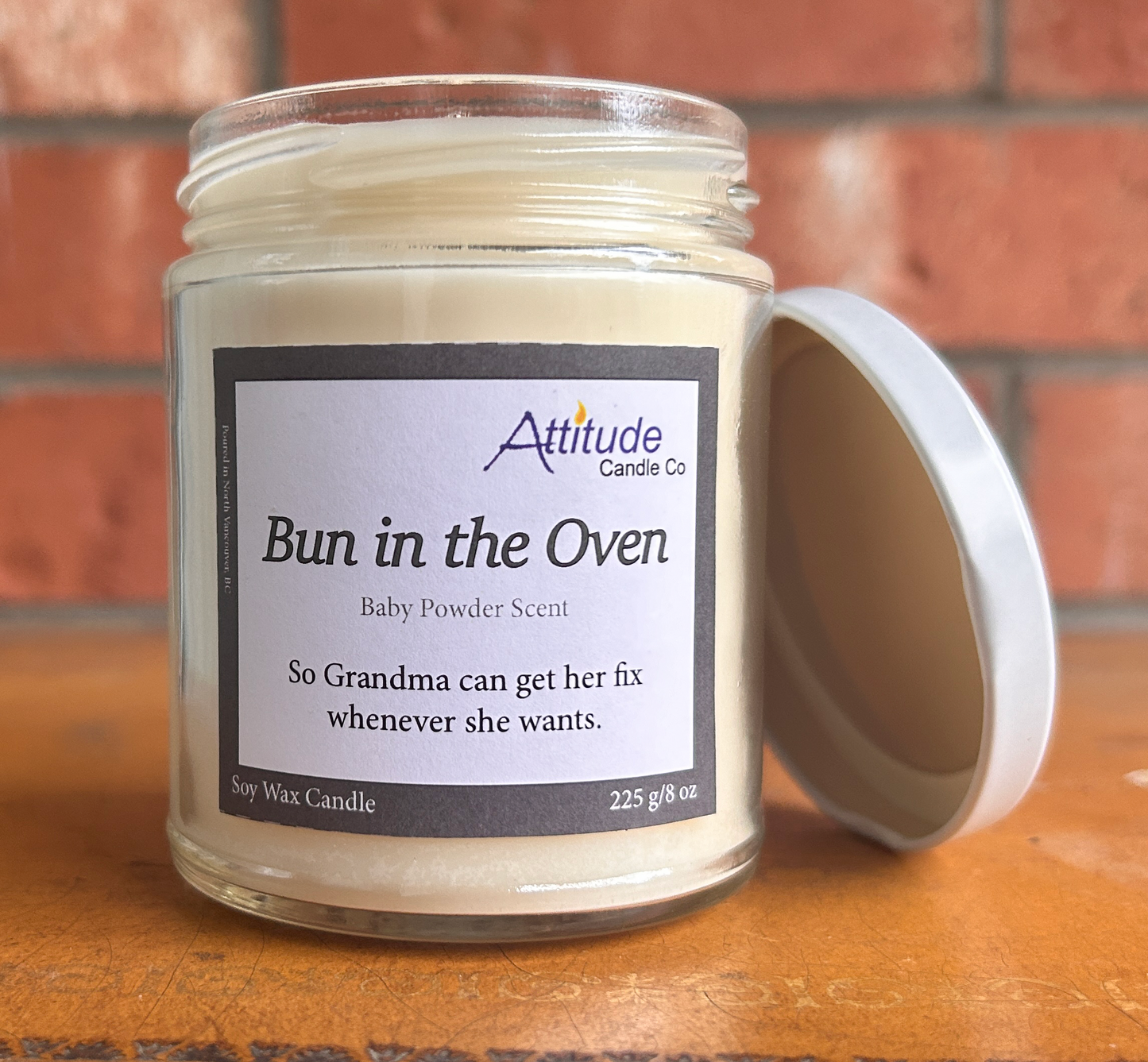 A picture of a candle in a jar with its lid off. The label says "Bun in the Oven", "Baby Powder Scent", and "So Grandma can get her fix whenever she wants". The label has an Attitude Candle Co logo in the top right corner. The candle is sitting on a brown table with a brick background behind.