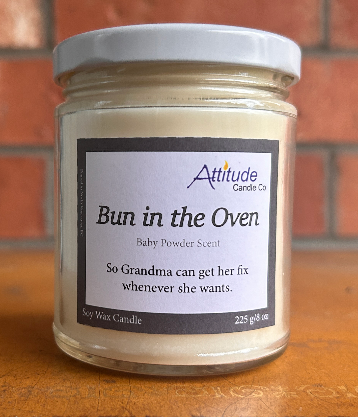 A picture of a candle in a jar with its lid on. The label says "Bun in the Oven", "Baby Powder Scent", and "So Grandma can get her fix whenever she wants". The label has an Attitude Candle Co logo in the top right corner. The candle is sitting on a brown table with a brick background behind.