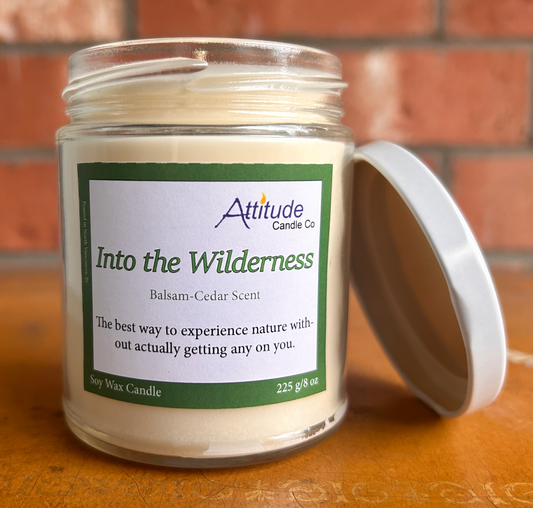 A picture of a candle in a jar with its lid off. The label says "Into the Wilderness", "Balsam-Cedar Scent", and "The best way to experience nature without actually getting any on you". The label has an Attitude Candle Co logo in the top right corner. The candle is sitting on a brown table with a brick background behind.