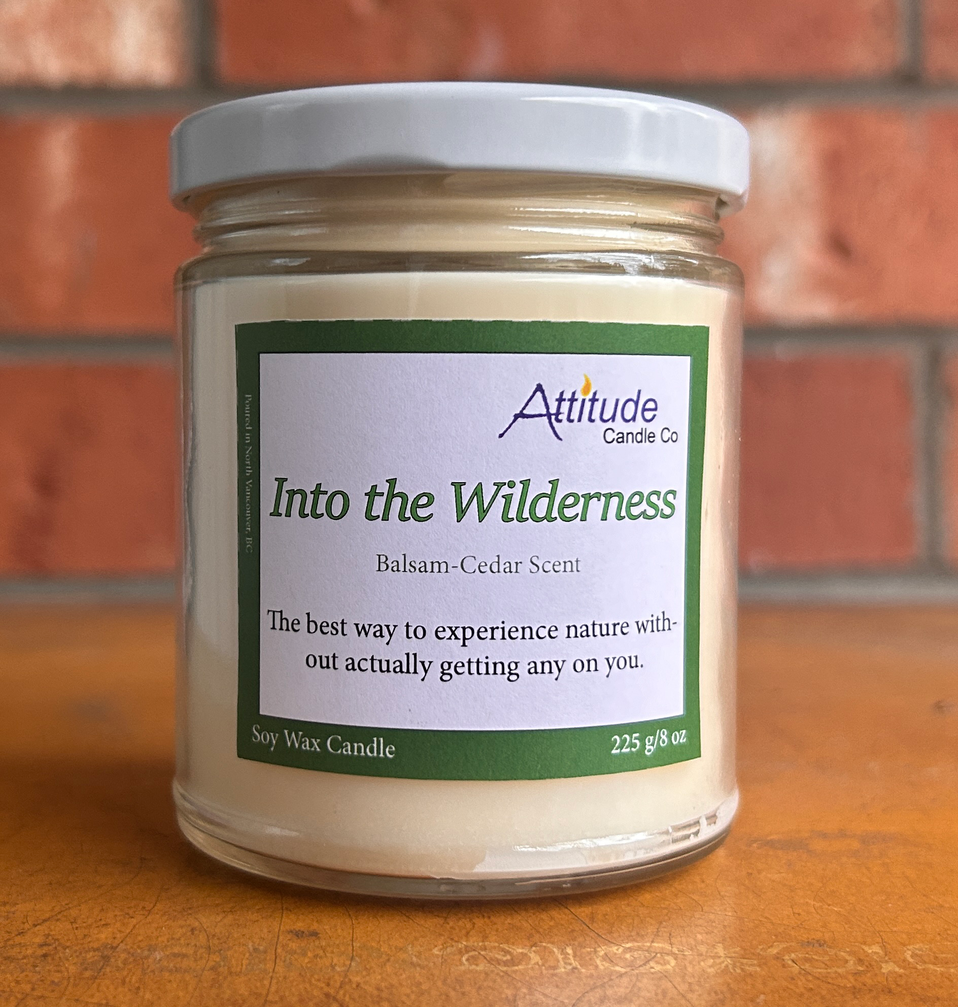 A picture of a candle in a jar with its lid on. The label says "Into the Wilderness", "Balsam-Cedar Scent", and "The best way to experience nature without actually getting any on you". The label has an Attitude Candle Co logo in the top right corner. The candle is sitting on a brown table with a brick background behind.