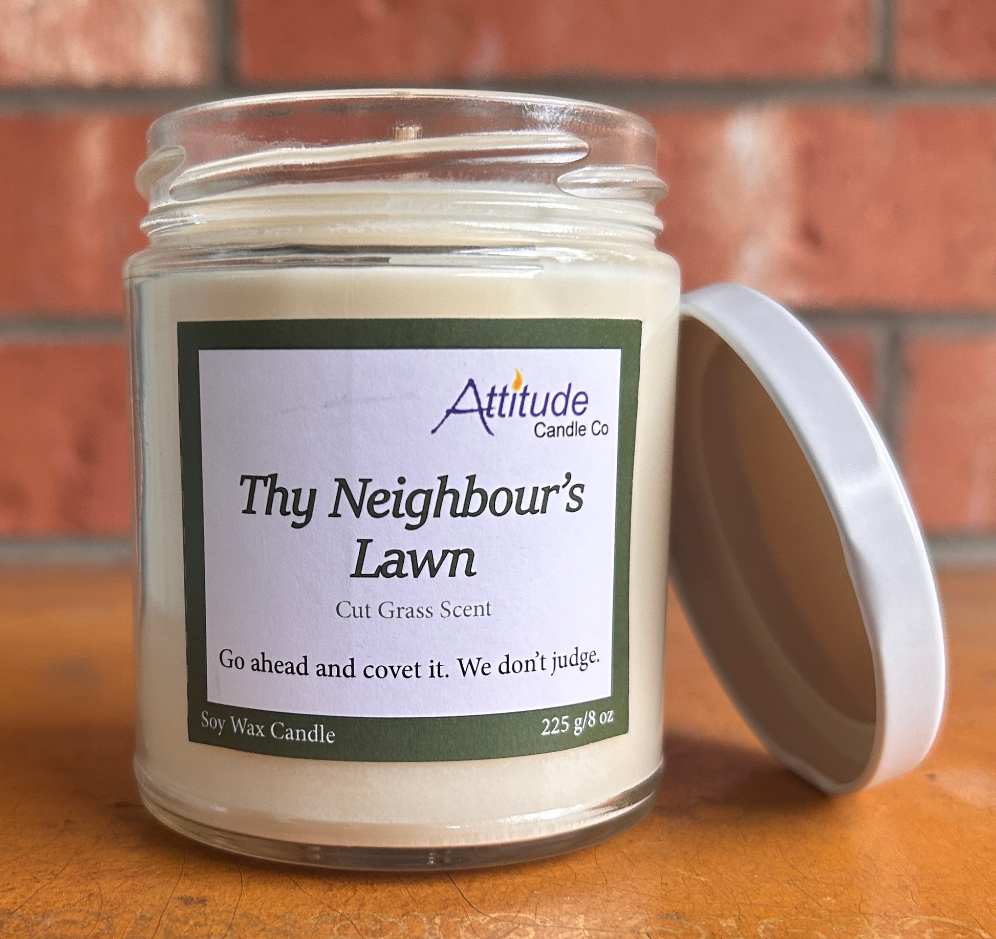 A picture of a candle in a jar with its lid off. The label says "Thy Neighbour's Lawn", "Cut Grass Scent", and "Go ahead and covet it. We don't judge". The label has an Attitude Candle Co logo in the top right corner. The candle is sitting on a brown table with a brick background behind.