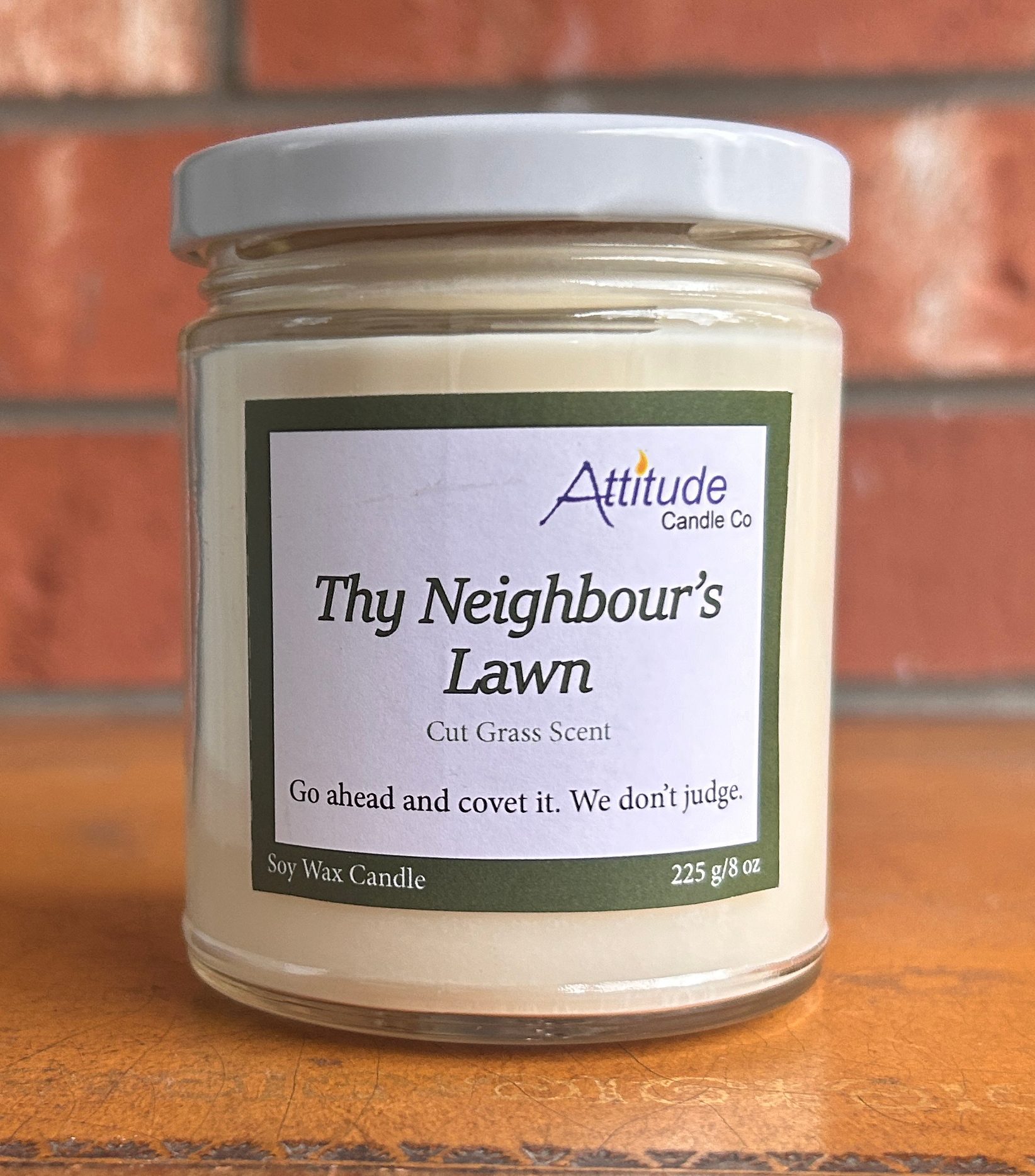 A picture of a candle in a jar with its lid on. The label says "Thy Neighbour's Lawn", "Cut Grass Scent", and "Go ahead and covet it. We don't judge". The label has an Attitude Candle Co logo in the top right corner. The candle is sitting on a brown table with a brick background behind.