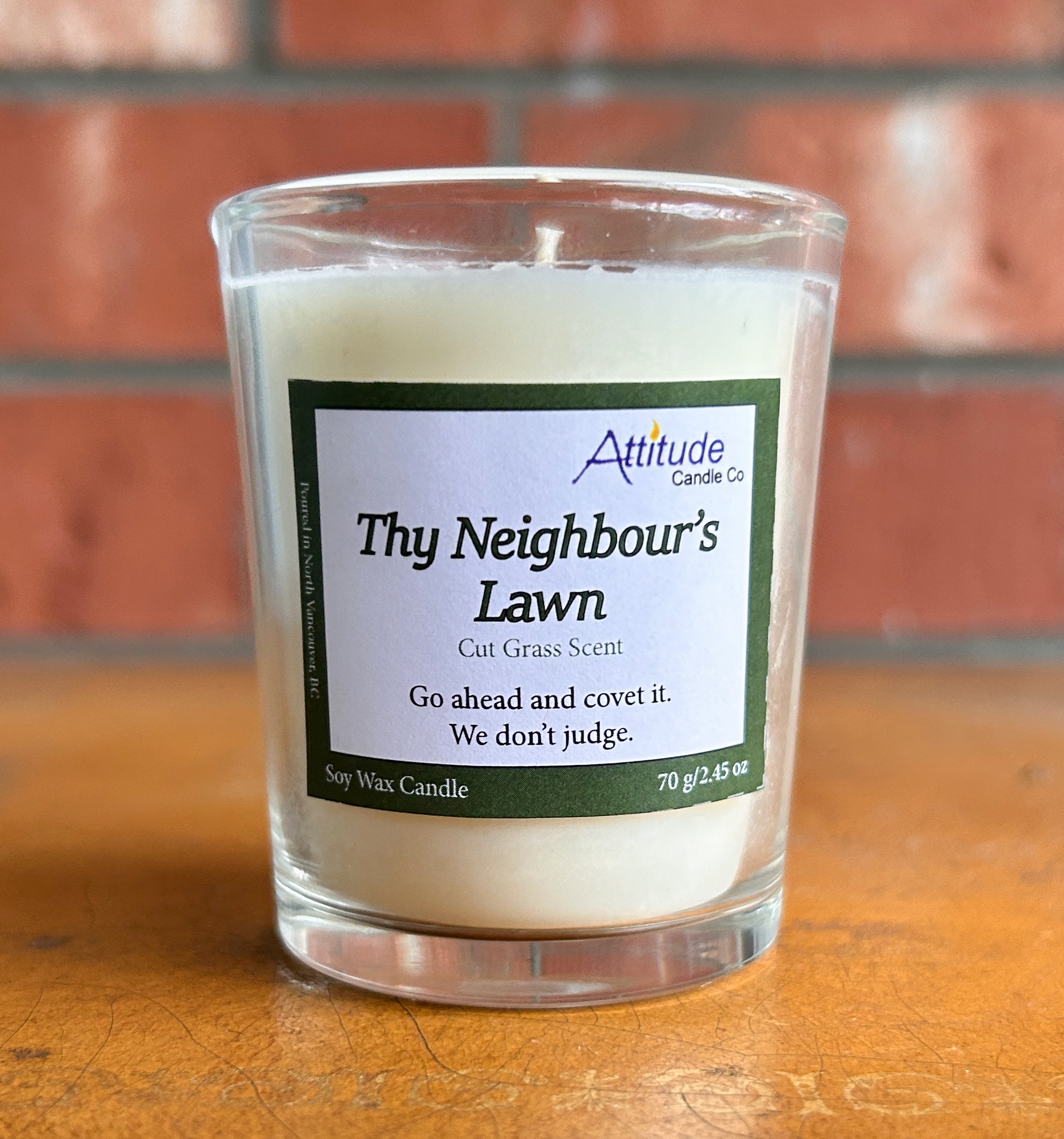 A picture of a votive candle in a jar. The label says "Thy Neighbour's Lawn", "Cut Grass Scent", and "Go ahead and covet it. We don't judge". The label has an Attitude Candle Co logo in the top right corner. The candle is sitting on a brown table with a brick background behind.