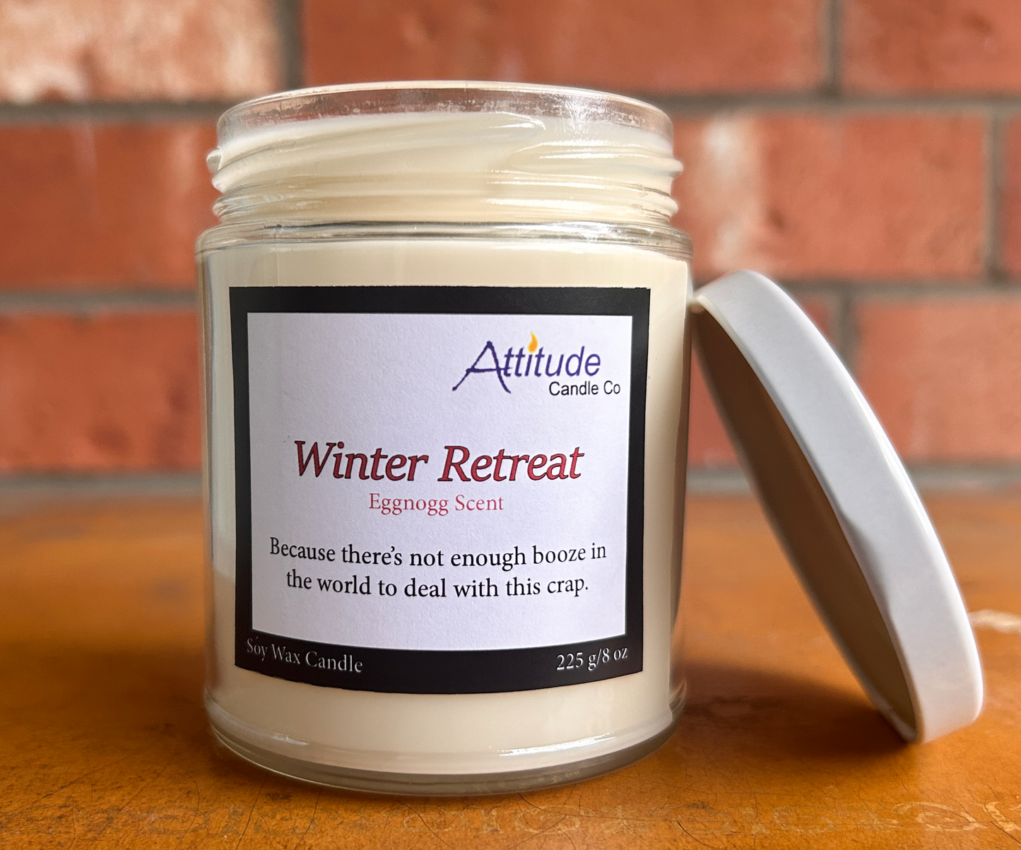 A picture of a candle in a jar with its lid off. The label says "Winter Retreat", "Eggnog Scent", and "Because there's not enough booze in the world to deal with this crap". The label has an Attitude Candle Co logo in the top right corner. The candle is sitting on a brown table with a brick background behind.