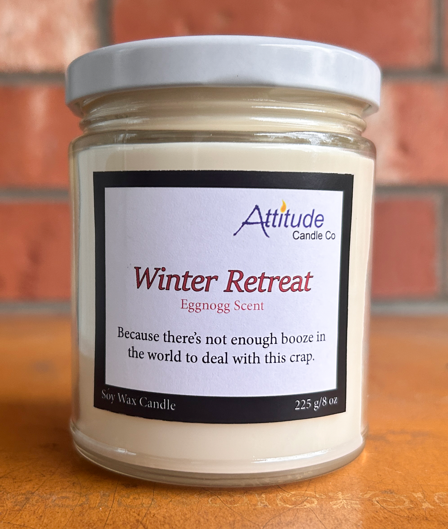 A picture of a candle in a jar with its lid on. The label says "Winter Retreat", "Eggnog Scent", and "Because there's not enough booze in the world to deal with this crap". The label has an Attitude Candle Co logo in the top right corner. The candle is sitting on a brown table with a brick background behind.