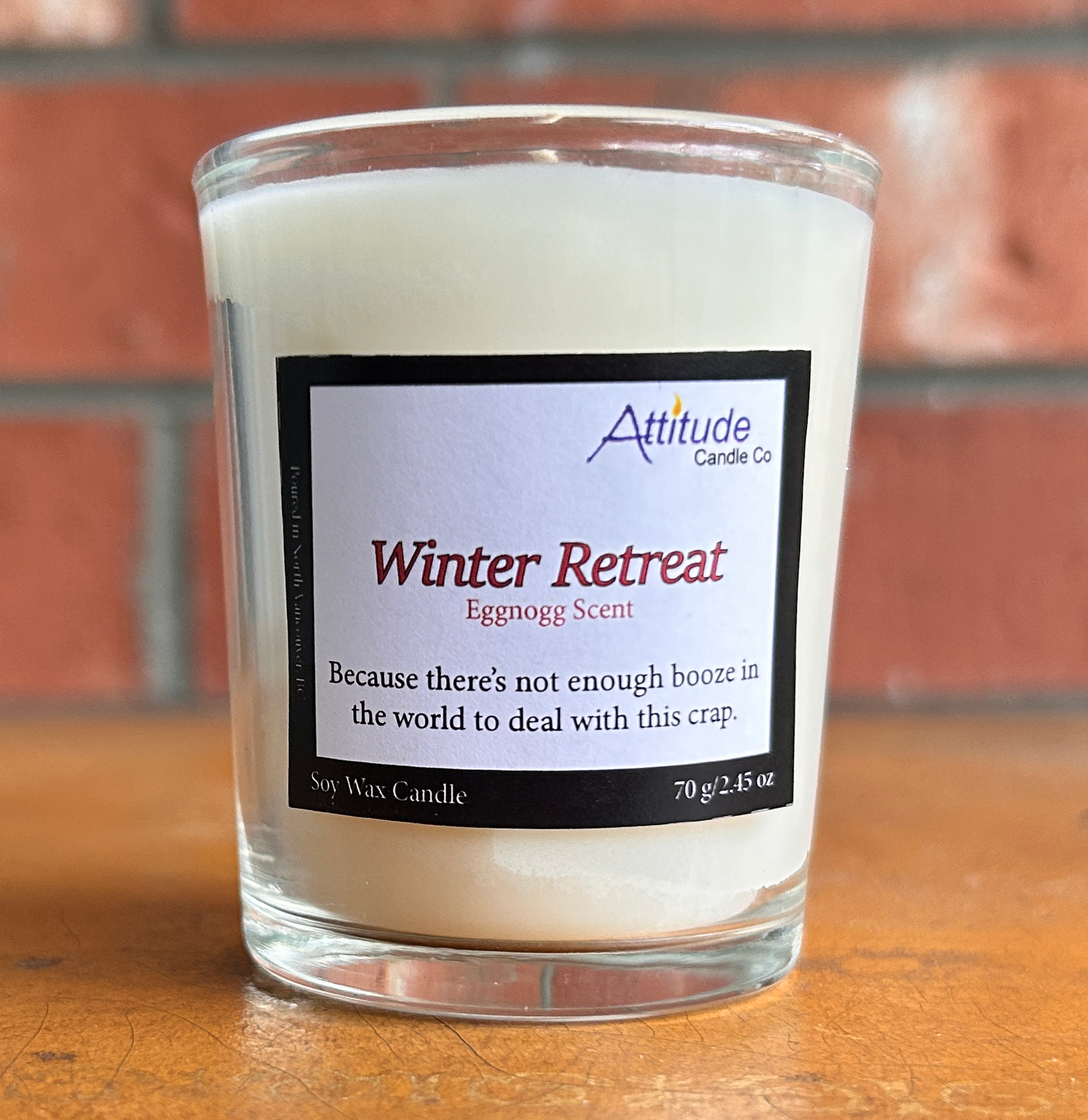 A picture of a votive candle in a jar. The label says "Winter Retreat", "Eggnog Scent", and "Because there's not enough booze in the world to deal with this crap". The label has an Attitude Candle Co logo in the top right corner. The candle is sitting on a brown table with a brick background behind.